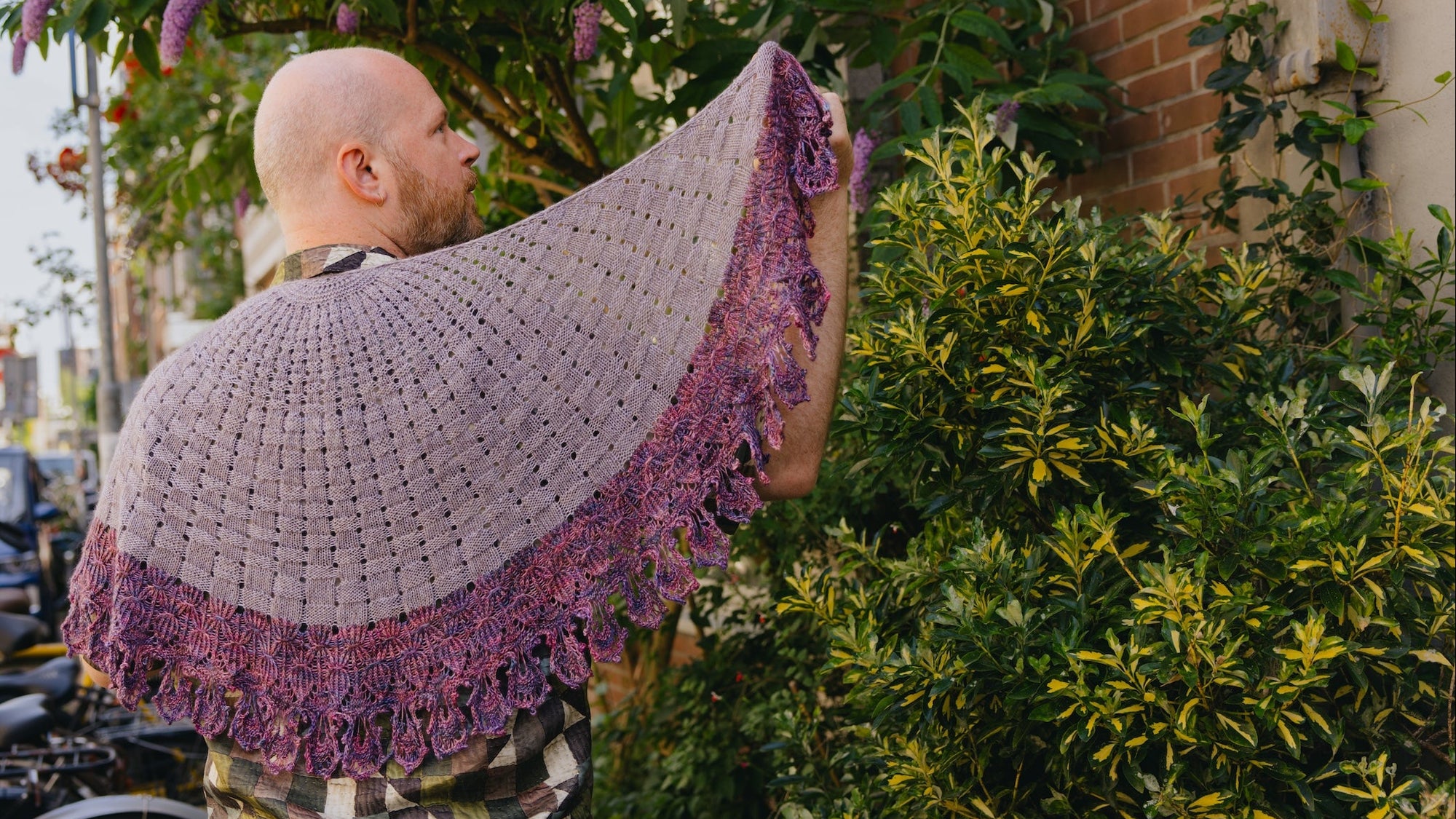 Stephen West in a Bouquet Basket Shawl Knitted pattern with Qing Fibre Yak Singles in purple in an outdoor setting. 